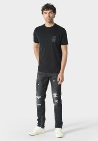 Černé bavlněné tričko s texturovaným logem na hrudi, spárované s ošuntělými tmavými džíny a bílými teniskami. Volný střih.