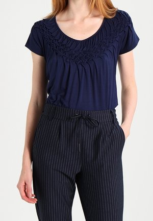 Femme portant un haut bleu marine à manches courtes avec un col texturé et un pantalon noir à fines rayures avec une taille à cordon, mains le long du corps.