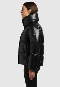 Veste matelassée noire avec un col montant, motif matelassé, poches zippées et à pression, avec un patch logo sur la manche gauche. Texture lisse et brillante.