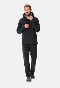Giacca nera impermeabile con cappuccio, chiusura con zip e polsini regolabili, abbinata a pantaloni neri e scarpe grigie. Texture liscia, senza motivi.