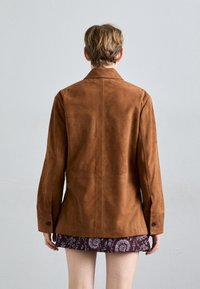 Άτομο με κοντά μαλλιά που φοράει ένα καφετί σ�ουέντ μπουφάν και μια μοτίβο φούστα, στέκεται μπροστά σε έναν απλό ανοιχτό γκρι τοίχο.