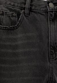 Gros plan sur un jean en denim noir avec une poche avant effilochée, des rivets en métal, une passants de ceinture et une fermeture boutonnée à la taille.