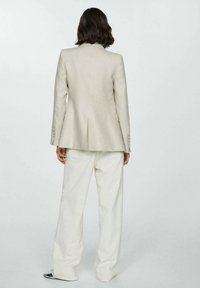 Blazer beige cintré avec une fente unique, deux poches et des épaules structurées. Associé à un pantalon blanc ample.