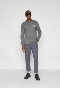 Camisa cinza de manga longa com gráfico na frente, combinada com calças cinza claras e sapatos pretos. O modelo usa óculos de sol pretos. Design simples e casual.