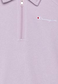 Světle fialový pulovr se zipovým límcem, vyrobený z měkké látky, s bílou vyšívanou logem a texturovaným pleteným vzorem.