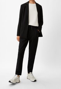 Black blazer over a white top, paired with tapered black trousers featuring a drawstring waist. Cream sneakers with textured accents.