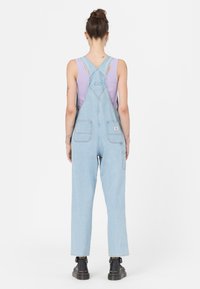 Salopette in denim azzurro chiaro con bretelle regolabili, due tasche posteriori e una vestibilità rilassata, indossata sopra una canotta lavanda. Scarpe nere completano il look.
