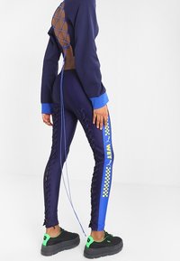 Granatowy sportowy top z siateczkowym tyłem, w zestawie z dopasowanymi granatowymi legginsami z sznurowaniem po bokach oraz niebieskimi panelami bocznymi z żółtym wzorem w kratkę.