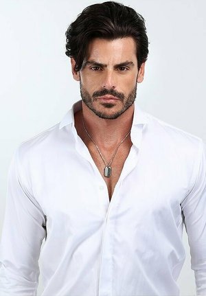 Camicia bianca con colletto, abbinata a una collana in argento con pendente a forma di dog tag. Capelli scuri pettinati all'indietro.