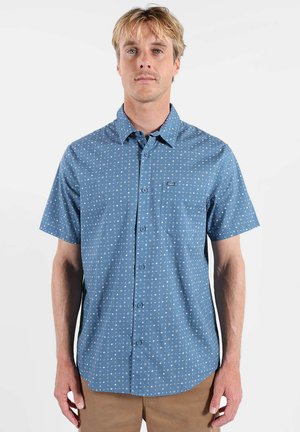 Homme portant une chemise à manches courtes bleu clair à boutons avec de petits motifs géométriques blancs et un pantalon beige, debout devant un fond blanc uni.
