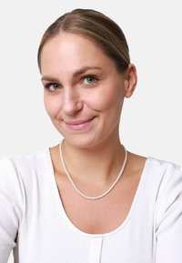Collier de perles blanches avec des perles de taille uniforme, porté par une femme dans un haut blanc côtelé, sur un fond gris uni.