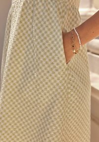 Robe à carreaux verts et blancs, en tissu texturé avec une poche, associée à des bracelets superposés en perles dorées et blanches.