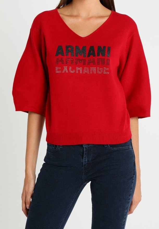 Femme portant un pull rouge à col en V avec des manches bouffantes et le texte "ARMANI" en strass noirs et rouges, associé à un jean bleu foncé.