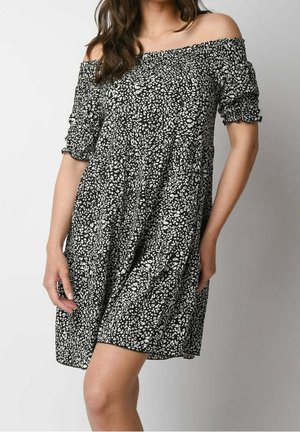 Kobieta ubrana w czarno-białą sukienkę z opuszczanymi ramionami, z półrękawkami i luźnym, zwiewnym krojem, stojąca na jednolitym tle.