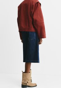 Veste courte couleur rouille, jupe en jean bleu foncé, et bottines beige en daim avec boucle. Des surfaces plates et texturées sont visibles.