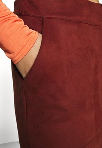 Jupe en daim rouge avec une texture lisse et une poche latérale. Gros plan sur la poche et la manche d'un vêtement orange. Ajustement soigné visible.