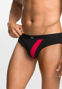 Pantaloni intimi neri in cotone con una striscia diagonale rossa; presentano una superficie liscia e un'etichetta con logo in vita. Progettati per comfort e vestibilità.