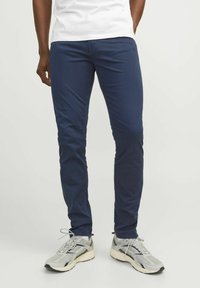 Námornícky modré chinos slim-fit vyrobené z elastickej bavlny, s plochým predným dielom a bočnými vreckami, v kombinácii s svetlošedými športovými topánkami.