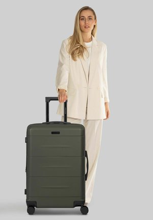 Grüner Hartschalenkoffer mit strukturierter Oberfläche, ausziehbarem schwarzen Griff und vier schwarzen Rollen, neben einer Person in beiger Kleidung stehend.