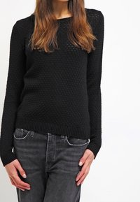 En kvinna som bär en svart strukturerad stickad tröja och mörkgrå jeans, stående med avslappnade händer vid sidorna mot en vit bakgrund.