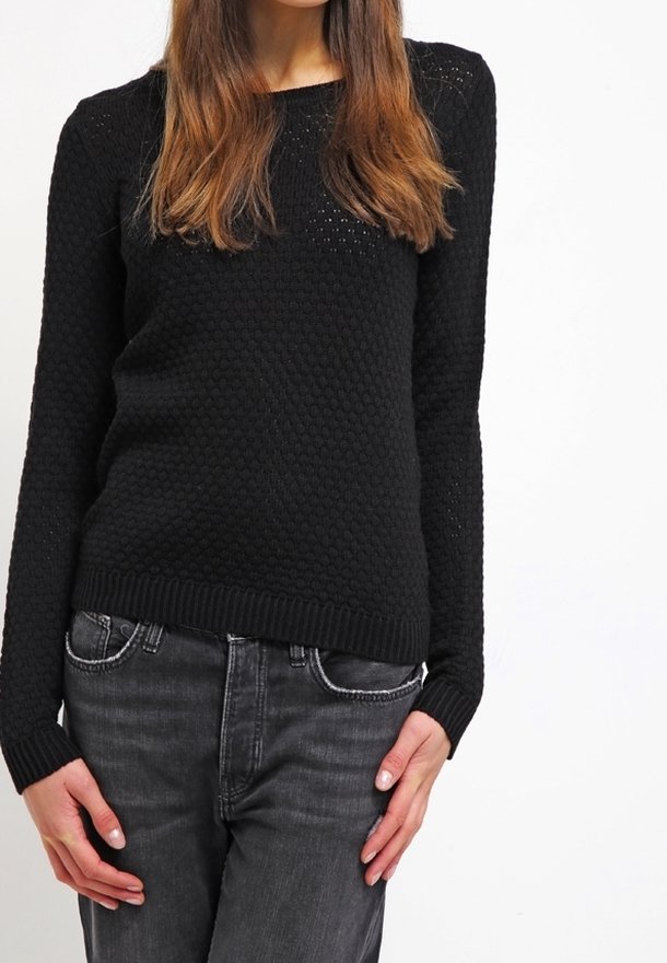 En kvinna som bär en svart strukturerad stickad tröja och mörkgrå jeans, stående med avslappnade händer vid sidorna mot en vit bakgrund.