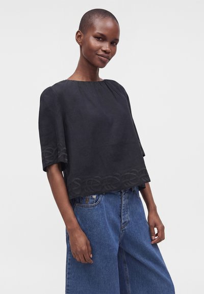 Femme aux cheveux courts portant un haut noir ample avec un ourlet brodé et un jean bleu large, debout devant un fond clair uni.