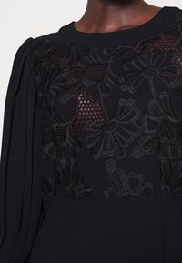 Černé šaty s texturovanou květinovou krajkovou aplikací na korzetu, s hladkým materiálem a strukturovanými balónovými rukávy.