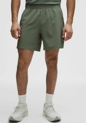 Mann trägt olivgrüne Sportschuhe, weiße Kniestrümpfe und weiße Laufschuhe, steht vor einfarbigem Hintergrund.