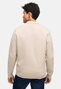 Beżowy sweter z teksturowanym splotem, posiada kołnierz, długie rękawy i prosty dół. Noszony na ciemnych dżinsach.