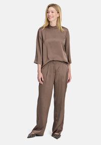 Top in satin marrone con scollo alto e maniche a tre quarti, abbinato a pantaloni in satin marrone plissettati. Scarpe a punta con borchie.