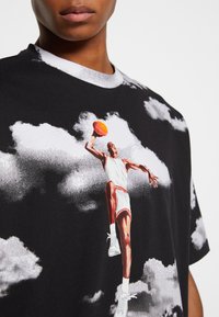 T-shirt en coton noir avec un imprimé graphique d'un joueur de basketball sautant sur un fond à motifs nuageux. Design à col rond.