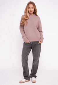 Kobieta o długich blond włosach, ubrana w fioletowy, miękki sweter, luźne ciemnoszare dżinsy i białe trampki, stojąca i uśmiechająca się.