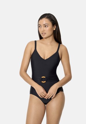 Mujer con cabello largo y oscuro, vestida con un traje de baño de una pieza negro con tirantes finos y un cinturón decorativo en la cintura, posando frente a un fondo liso.