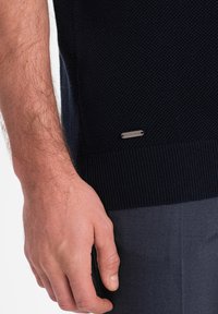 Pull en maille bleu marine avec un détail de bas ribbé. Étiquette métallique visible sur le côté inférieur. Une main repose au bord du pull.