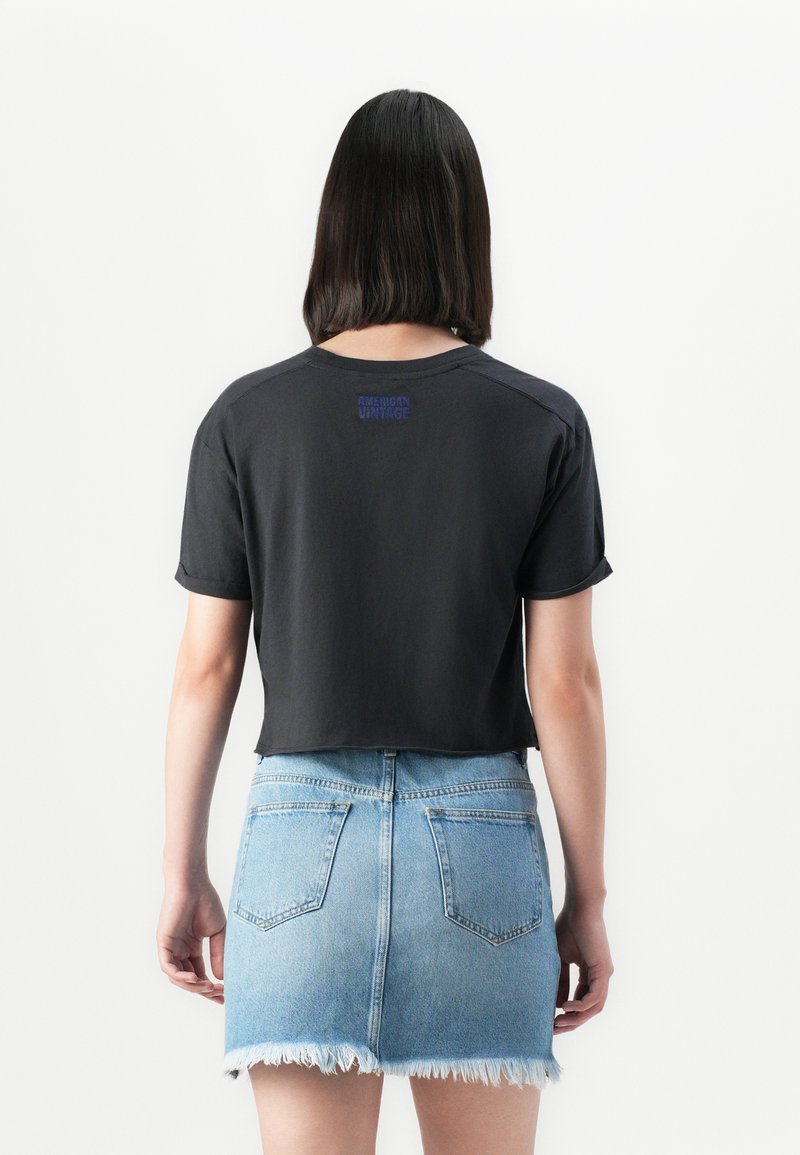 Pessoa com cabelo preto até aos ombros, vestindo uma t-shirt preta curta e uma saia mini de ganga azul clara desfiada, de costas.