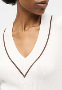 Κοντινό πλάνο ενός ατόμου που φοράει λευκό πλεκτό πουλόβερ με V λαιμόκοψη και λεπτή καφέ ρίγα κατά μήκος του λαιμού.