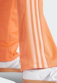 Orange track pants with pink side stripes, made of textured fabric, paired with white shoes featuring a gum sole and minimal design.