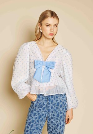 Szőke nő áttetsző fehér blúzban, világoskék pöttyökkel és nagy kék masnival, texturált kék mintás nadrággal párosítva.