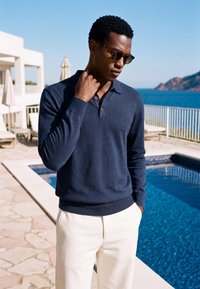 Homme portant des lunettes de soleil, un polo bleu marine à manches longues et un pantalon blanc, debout près d'une piscine avec l'océan et des montagnes en arrière-plan.