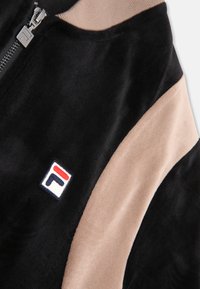 Mjukt svart och beige jacka med dragkedja och ett litet rektangulärt logotyg i röd, vit och marinblå på bröstet.