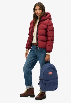 Mujer con chaqueta acolchada roja, vaqueros azules con puños, botas marrones y que sostiene una mochila azul con un parche de la bandera estadounidense.