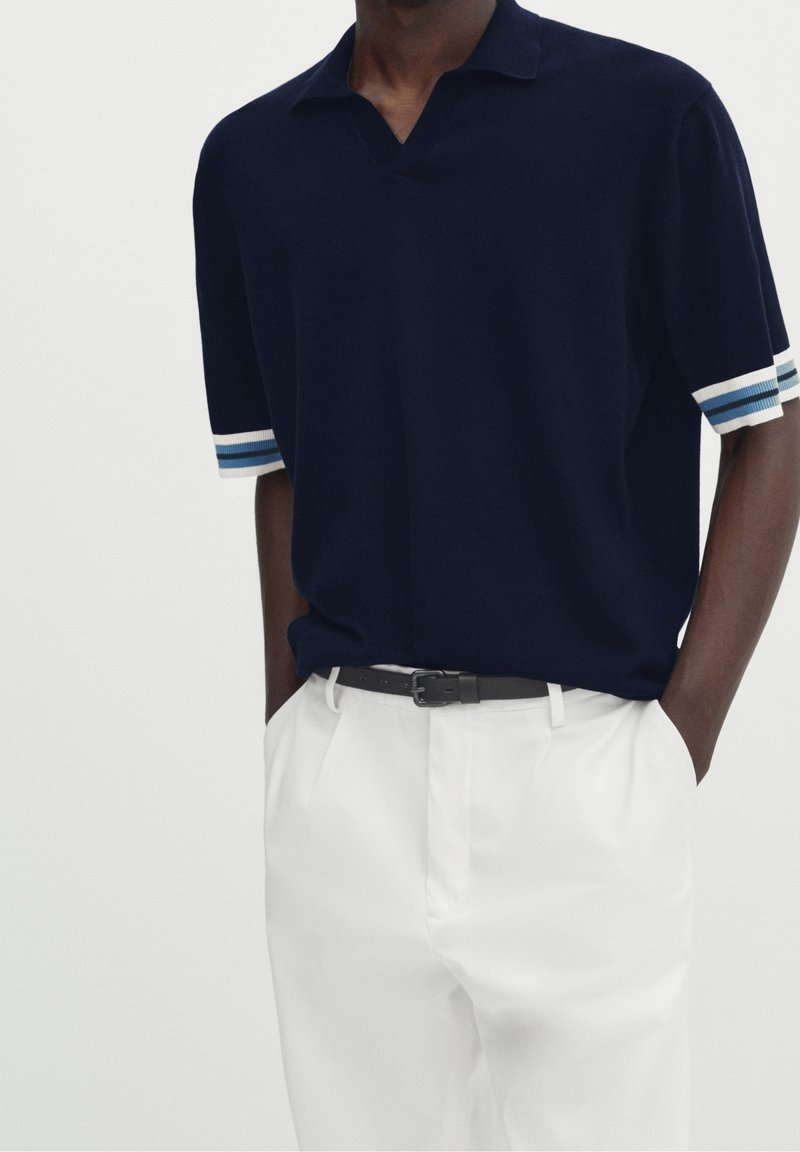 Homme portant un polo à manches courtes bleu marine, avec des poignets rayés bleus et blancs, rentré dans un pantalon blanc à plis, avec une ceinture noire.