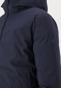 Mørkeblå vandtæt jakke med hætte, der har lynlås foran, glat tekstur og et cirkulært logo-patch på venstre ærme.