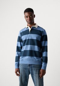 Polo à manches longues rayé bleu marine et bleu clair avec un col blanc. Le tissu est lisse et la coupe semble décontractée. Porté avec un jean en denim clair.