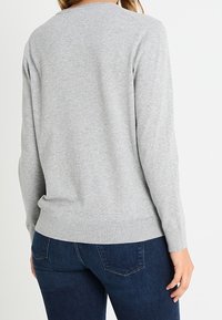 Šedý svetr s dlouhými rukávy, kulatým výstřihem a žebrovaným lemem, vyrobený z měkké látky. Nošený přes tmavé džíny, představující jednoduchý design.