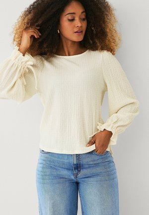 Kvinde med krøllet hår iført en tekstureret cremefarvet bluse med puffede ærmer og lyseblå jeans, poserer med den ene hånd i lommen.