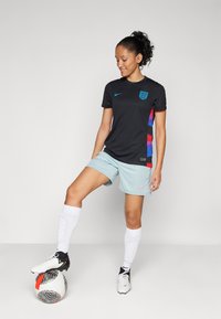 Black Nike sports jersey with blue accents and England crest, paired with mint green shorts. White knee-high socks and black soccer cleats complete the outfit.