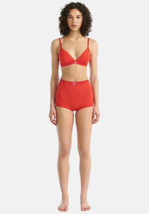 Frau mit lockigem Haar, die ein passendes rotes, strukturiertes BH- und hoch tailliertes Shorts-Set mit Logo-Akzenten trägt, barfuß vor einem weißen Hintergrund steht.