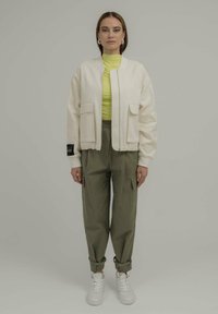 Veste bomber blanche avec deux poches avant, associée �à un haut côtelé jaune vif, un pantalon vert olive et des baskets blanches.