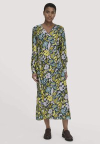 Eine Frau trägt ein langärmliges, V-Ausschnitt-Midi-Kleid mit einem bunten Blumenmuster und dunklen braunen Loafers, stehen vor einem einfarbigen Hintergrund.